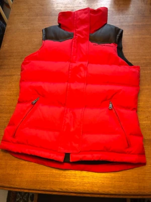 Ny röd dunväst Peak Performance. Stl S. - Varm röd dunväst från Peak Performance med svarta axelpartier i skinnimitation och dragkedjeförsedda fickor. Ny utan prislapp. Riktigt rejält med dun i denna väst samt kraftig tyg. (Kolla bild nr 5, den står helt själv utan stöd😂). 