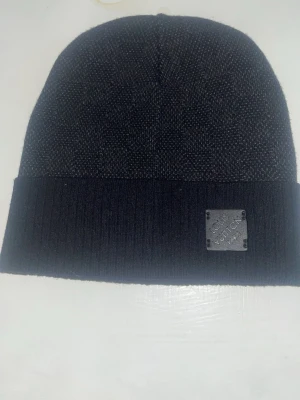  En svarta  mössa  - Snygg svart mössa   med diskret rutmönster och ribbad uppvikt kant. Märkets metallbricka sitter framtill och ger en exklusiv touch. Perfekt accessoar för dig som gillar stilrena och lyxiga detaljer. Materialet känns mjukt och värmande.