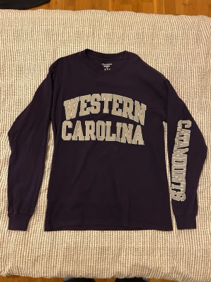 Långärmad lila Champion t-shirt - Långärmad t-shirt från Champion i mörklila med trycket 'Western Carolina' på bröstet och 'Catamounts' på ena ärmen. Den är köpt på Beyond Retro och är därför vintage från USA. Kan vara 90 tal. Inget tryck på ryggen. Jag är 174 och den sitter bra på mig, lagom stor.