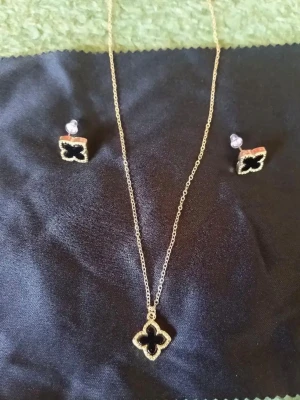 Guldigt halsband och örhängen set - Snyggt smyckeset med halsband och matchande örhängen i guldton. Båda har fyrklöverformade hängen med svart detalj i mitten. Perfekt för dig som vill ha en stilren och trendig accessoar med lite lyxig känsla.