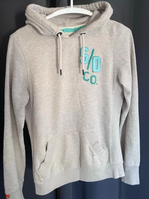 Superdry Hoodie - Snygg grå hoodie från Superdry med turkosa broderade detaljer fram och på huvan. Klassisk känguruficka, dragsko i huvan och mjukt material. Perfekt för en avslappnad och trendig look.