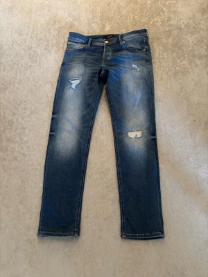 Blå slitna slim jeans från Jack and Jones  - Säljer ett par blå slim fit jeans från Jack and Jones med snygga slitningar och lätt tvättade detaljer. Jeansen har klassisk femficksmodell, knappgylf och kontrastsömmar. Perfekta för en trendig och avslappnad look.