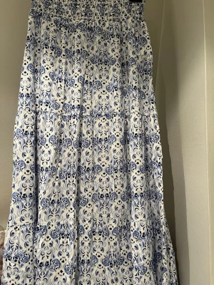 Vit och blå långkjol med mönster - Säljer en långkjol i vitt med blått, detaljerat mönster. Kjolen är luftig och perfekt för varma dagar. Tyget känns lätt och svalt, och mönstret ger en härlig sommarkänsla. Endast testad.