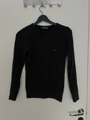 Svart kabelstickad tröja Tommy Hilfiger - Svart kabelstickad tröja från Tommy Hilfiger med rund halsringning och diskret logga på bröstet. Tröjan har långa ärmar och ribbade muddar vid ärmslut och nederkant. Perfekt för en stilren och klassisk look.