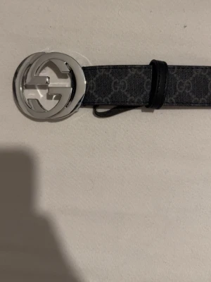 Svart Gucci bälte med silverdetaljer - Snyggt svart bälte från Gucci med klassiskt GG-mönster och stor silverfärgad GG-spänne. Bältet har en stilren design och är perfekt för att lyfta vilken outfit som helst. Kommer med originalask.