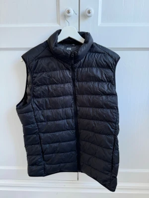 Svart dunväst från Uniqlo - Stilren svart dunväst från Uniqlo med hög krage och dragkedja framtill. Västen har en quiltad design med horisontella sömmar och är lätt vadderad för extra värme. Perfekt att slänga över en hoodie eller tröja för en casual look.