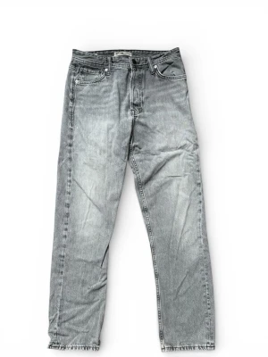 Jack & Jones jeans - Säljer ett par grå jeans från Jack & Jones med rak passform och klassisk femficksdesign. Jeansen har en snygg vintage-look med ljusa slitningar och gråa kanter vid benslut. Tillverkade i bomull för en skön känsla och stilren vibe.