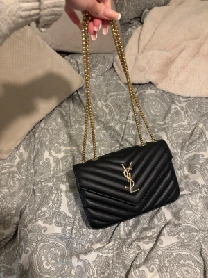 Svart YSL handväska med guldkedja - Säljer en svart handväska från Yves Saint Laurent med quiltat mönster och ikonisk YSL-logga i guld framtill. Väskan har en elegant guldfärgad kedjerem och är tillverkad i slätt läder. Perfekt accessoar för att lyfta din outfit.