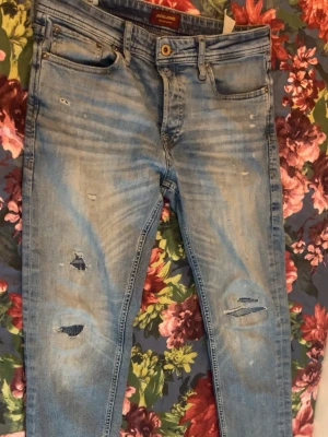 Ljusblå slitna jeans Jack & Jones - Säljer ett par ljusblå jeans från Jack & Jones med slitningar och hål på knäna för en trendig, avslappnad look. Klassisk femficksmodell med knappgylf och skinny fit. Perfekta för dig som gillar streetstyle och vill ha en cool vibe.