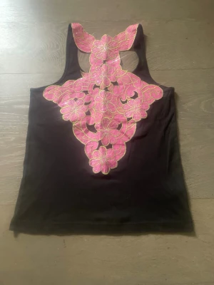 Y2k Svart linnetopp med rosa spetsdetalj - Snygg svart y2k 90' linnetopp med unik rygg i rosa spets och broderade blommor. Toppen har ärmlös design och ett stort blommönster i rosa med guldkant som täcker hela ryggen. Perfekt för dig som vill sticka ut med coola detaljer.