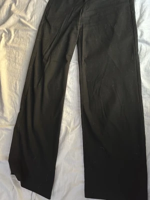 Svarta vida kostymbyxor - Säljer ett par svarta kostymbyxor med vid passform och hög midja. Byxorna har raka ben och är tillverkade i ett mjukt, vävt material som ger en snygg siluett. Perfekta för dig som gillar en stilren och trendig look. En söm har gått upp vid höger ankel. 