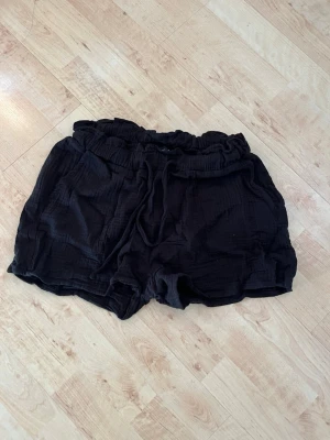 Svarta shorts med snörning - Säljer ett par svarta shorts med resår i midjan och snörning framtill. De är tillverkade i ett mjukt, lite krinklat bomullstyg och har en avslappnad passform. Perfekta för varma dagar och enkel att matcha med olika toppar.