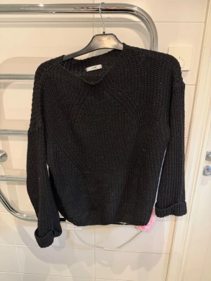 Svart stickad tröja från Hailys - Snygg svart stickad tröja från Hailys med v-ringning och grovstickad struktur. Tröjan har långa ärmar och en avslappnad passform, perfekt för lager på lager. Materialet känns mjukt och bekvämt, och den ribbade kanten ger en extra touch. Skulle inte säga att passformen är XL, passar mer S/M