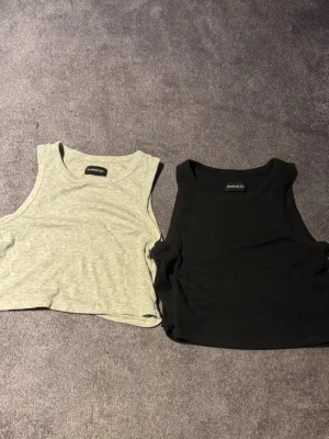 2 croppade ribbade linnen Even&Odd - Säljer två croppade linnen från Even&Odd, ett i ljusgrått och ett i svart. Båda är ribbade och ärmlösa med rund halsringning. Perfekta för sommaren och enkla att matcha till olika outfits.