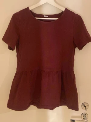 Vinröd topp från Lindex - Vinröd topp från Lindex med rund halsringning och korta ärmar. Modellen har en lätt utställd nederdel som ger en söt siluett. Tillverkad i mjukt bomullstyg, perfekt för dig som gillar enkel men snygg stil.