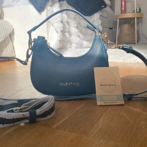 Blå axelväska från Valentino - Helt ny marin blå axelväska från Valentino med guldfärgade detaljer och dragkedja. Väskan har en stilren bågform och kommer med två axelband, ett smalare i blått skinn och ett bredare i blått och vitt tyg med logga. 💙