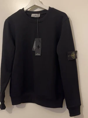 Svart sweatshirt från Stone Island - Svart sweatshirt från Stone Island med rund hals och ribbade muddar. Klassisk patch med logga på vänster ärm som går att ta av. Tillverkad i mjuk bomull för skön känsla och clean look. Perfekt för dig som gillar streetwear och enkel stil.