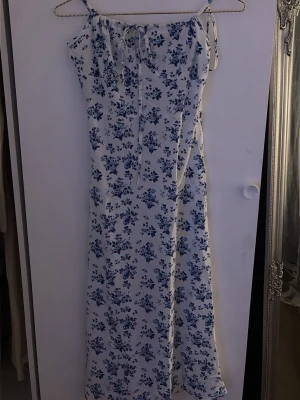 Vit långklänning med blå blommor - Säljer en vit långklänning med blått blommönster och tunna axelband. Klänningen har en smickrande rak passform och knytband framtill. Perfekt för sommarens alla tillfällen och ger en fräsch och romantisk look.