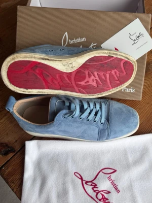 Blåa sneakers från Christian Louboutin som nya - Snygga blåa sneakers i mocka från Christian Louboutin med klassisk röd sula och vita kanter. Skorna har rund tå, matchande blå skosnören och diskret branding på sulan. Perfekta för dig som vill ha något exklusivt och stilrent.