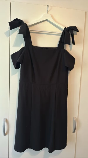 Svart klänning med rosettband - Stilren svart klänning från Lindex med axelband som knyts i stora rosetter. Klänningen har en rak passform, kort längd och är tillverkad i ett mjukt polyestertyg. Perfekt för dig som gillar en enkel men trendig look.