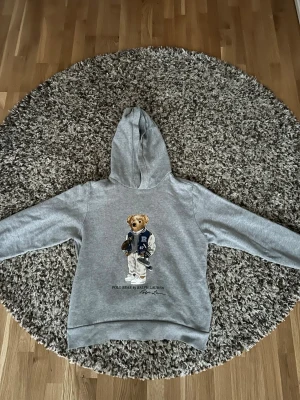 Grå hoodie Polo Ralph Lauren  - Säljer en grå hoodie från Polo Ralph Lauren. På ryggen finns ett stort tryck av den ikoniska björnen i collegekläder. Hoodien har huva och är gjord i mjukt bomullsmaterial. Perfekt för en chill och avslappnad stil.