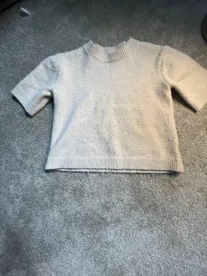 Beige stickad kortärmad tröja, ganska så noppig. - Säljer en beige stickad tröja med korta ärmar och ribbad kant vid hals och nederdel. Tröjan har en boxig passform och är gjord i ett mjukt, strukturerat material. Perfekt för lager på lager och enkel att matcha med olika outfits.