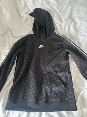 Svart Nike hoodie med logomönster - Snygg svart hoodie från Nike med heltryckt logomönster och vit Nike-logga på bröstet. Hoodien har huva, magficka och vita detaljer längs ärmarna. Tillverkad i mjukt material som känns skönt mot huden. Perfekt för dig som gillar streetwear och sportig stil.