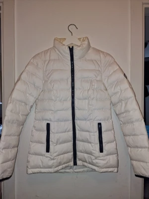 Vit pufferjacka från Tommy Hilfiger - Snygg vit pufferjacka från Tommy Hilfiger med svart dragkedja och svarta detaljer vid fickorna. Jackan har hög krage och ett broderat Tommy Hilfiger-märke på ärmen. Perfekt för en stilren look, lite missfärgning i kragen på insidan pga av smink därav lågt pris. Syns däremot inte när man har på sig den 🤍