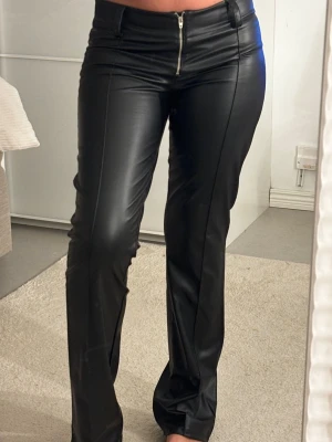 Svarta skinnbyxor  - Säljer ett par svarta byxor i skinn med låg midja och raka ben. Byxorna har en synlig dragkedja framtill, bälteshällor och söm längs benen för en snygg look. Byxan är i storlek 170💞