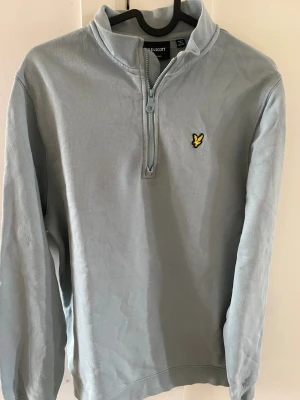 Ljusblå halvzip från Lyle & Scott - Ljusblå tröja från Lyle & Scott med halv dragkedja och klassisk gul logga på bröstet. Tröjan har hög krage och långa ärmar, perfekt för lager på lager. Materialet är mjukt och bekvämt, passar till många olika stilar.