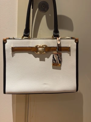 Vit handväska från Aldo med gulddetaljer - Stilren vit handväska från Aldo med svarta handtag och kanter, guldfärgade metalldetaljer och en brun dekorationsrem på framsidan. Väskan har ett ormmönstrat hänge och rektangulär form. Perfekt för dig som gillar klassisk men trendig design.