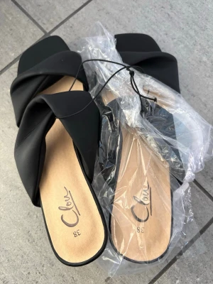 Svarta slip-in sandaler från Clou med klack nya - Stilrena svarta slip-in sandaler från Clou med breda korslagda remmar i konstläder och fyrkantig tå. Sandalerna har en låg klack och en beige innersula med logga. Perfekta för en trendig och avslappnad look. Oanvända. Små i storlek skulle passa 36-37 gissar jag på