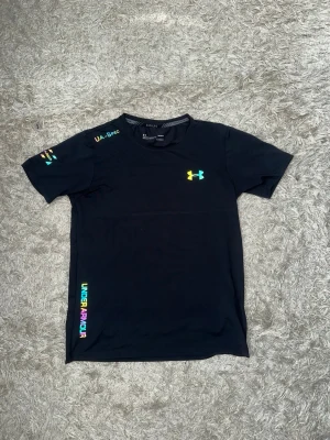 Svart Under Armour t-shirt (reflex) - Svart t-shirt från Under Armour med färgglada loggor och textdetaljer på bröstet, ärmen och sidan. Tillverkad i ett lätt och sportigt material som passar perfekt till träning. Klassisk passform med rund hals och korta ärmar. Hela settet finns i profilen som en egen annons