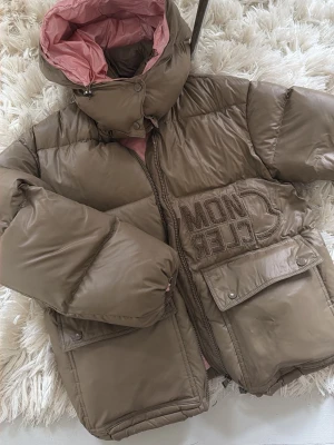 grön pufferjacka från Moncler - Snygg grön pufferjacka från Moncler med stor broderad logga på bröstet. Jackan har två stora fickor med lock och tryckknappar, hög krage och huva med rosa foder. Materialet är glansigt och ger en lyxig känsla. Perfekt för kalla vinterdagar.