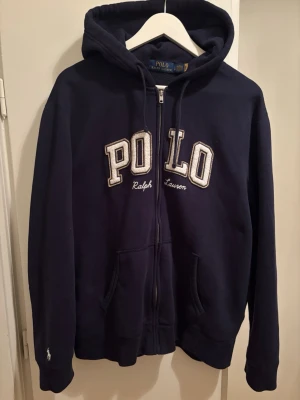 Marinblå hoodie från Polo Ralph Lauren - Mörkblå hoodie med dragkedja från Polo Ralph Lauren. Stor POLO-logga i vitt och grått broderad på bröstet och liten logga på ärmen. Utrustad med huva och två fickor framtill. Klassisk och stilren design i mjukt material.