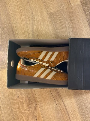 Adidas Wales Bonner bruna sneakers - Snygga sneakers från Adidas x Wales Bonner i brunt med vit sula och vita detaljer. Skorna har klassiska tre ränder på sidan, guldfärgade öljetter och croc-mönstrad ovandel i glansigt material. Perfekt för dig som vill sticka ut med en unik och trendig look.
