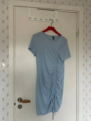 Ljusblå klänning med rynk från H&M - Kortärmad ljusblå klänning från H&M med rund halsringning och snygg rynkdetalj på ena sidan som ger en cool draperad effekt. Klänningen är enkel men trendig och har en justerbar dragsko nedtill. Perfekt för dig som gillar stilrena plagg med en twist.