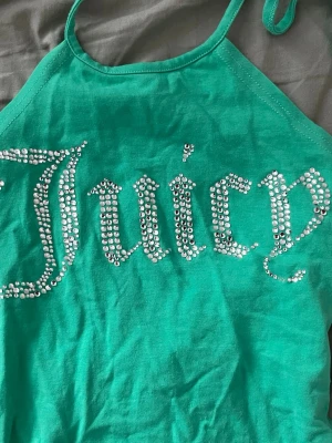 Grön Juicy Couture linnetopp - Snygg grön linnetopp från Juicy Couture med glittrande strassdetaljer som stavar 'Juicy' på framsidan. Toppen har tunna axelband som knyts i nacken och är perfekt för en trendig sommarlook.