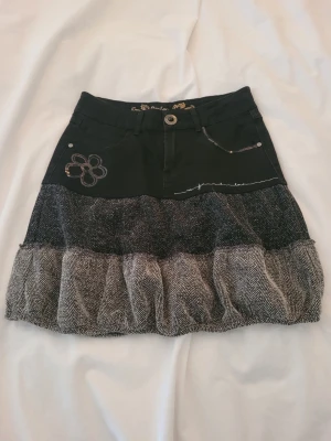 Svart och grå volangkjol från Desigual - Kort kjol från Desigual i svart och grått med volang och broderade detaljer som blomma och hjärta. Kjolen har fickor fram och bak samt dekorativa sömmar. Materialmix med struktur och mönstrad nederdel ger en unik look.