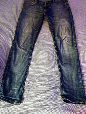 Nudie blå slitna jeans - Säljer ett par blå jeans med snyggt slitna detaljer och raka ben. Jeansen har klassisk femficksdesign och kontrastsömmar. Materialet är denim i bomull och de har en cool tvättad look som passar till det mesta.