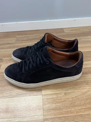 Blå mockasneakers med vit sula - Stilrena blå sneakers i mocka med vita sulor och brunt innerfoder i läder. Klassisk låg modell med blå skosnören och enkel design som passar till det mesta. Perfekta för dig som gillar en clean och tidlös look.