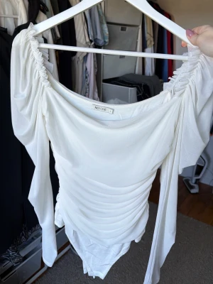 Vit off shoulder body från NLY One - Trendig vit body från NLY One med off shoulder-design och långa, puffiga ärmar. Materialet är stretchigt och lätt, med rynkade detaljer längs sidorna för en snygg siluett. Perfekt för dig som vill ha en stilren och modern look.