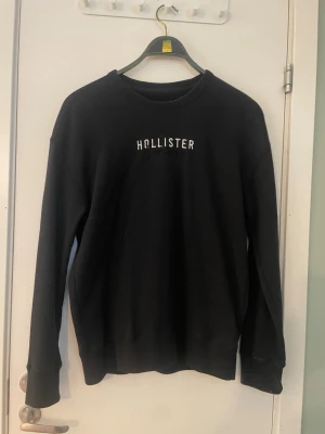 Svart sweatshirt från Hollister - Klassisk svart sweatshirt från Hollister med vit broderad logga på bröstet. Tröjan har rund halsringning, långa ärmar och ribbade muddar vid ärmslut och nederkant. Perfekt för en avslappnad och stilren look.