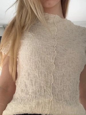 Ribbad topp med vågig söm - Trendig beige topp med ribbad struktur och vågig söm framtill och baktill. Kortärmad modell med lätt transparent material och hög hals. Står ej storlek men passar som Xs/S