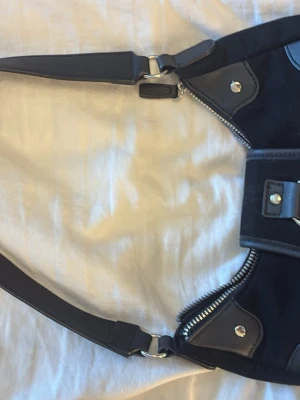 Svart axelväska med silverdetaljer - Cool svart axelväska med justerbar rem, silverfärgade dragkedjor och nitar. Väskan har en unik form och detaljer i både tyg och konstläder. Perfekt för dig som gillar edgy accessoarer och vill sticka ut.