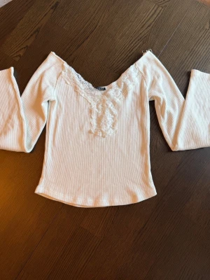 Vit ribbad topp med spetsdetaljer - Säljer en söt vit ribbad topp med lång ärm och v-ringning dekorerad med spets. Toppen har en feminin och mjuk look, perfekt för dig som gillar romantiska detaljer. Materialet känns stretchigt och bekvämt.