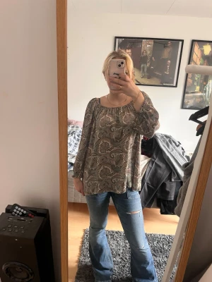Mönstrad paisleyblus med vida ärmar - Säljer en lös paisleymönstrad blus i beige och bruna toner. Blusen har vida långa ärmar med resår vid ärmslut och en rundad halsringning. Perfekt för dig som gillar boho-stil och vill ha något luftigt och bekvämt.
