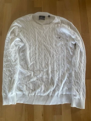 Vit kabelstickad tröja från GANT - Säljer en klassisk vit kabelstickad tröja från GANT med rund halsringning och ribbade muddar. Tröjan har GANT-logga och sköld broderad på bröstet. Perfekt för dig som gillar stilrena och tidlösa plagg i garderoben. 