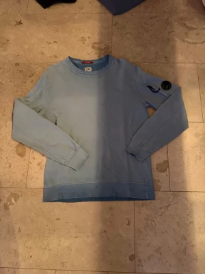 Ljusblå sweatshirt C.P. Company - Säljer en ljusblå sweatshirt från C.P. Company med rund halsringning och lång ärm. Tröjan har den ikoniska svarta linsen på ärmen och en liten ficka med dragkedja. Perfekt för dig som gillar streetwear och snygga detaljer.