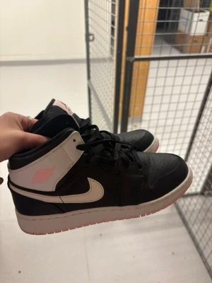 Air Jordan 1 Mid svart/rosa - Säljer ett par Air Jordan 1 Mid sneakers i svart och vitt med rosa detaljer. Skorna har klassisk hög siluett, svart läderöverdel, vita swoosh och rosa logga samt rosa detaljer på plösen och yttersulan. Perfekta för dig som gillar streetwear och snygga detaljer.